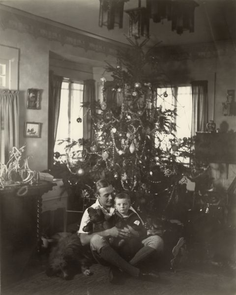 Silent film actor Wallace Reid sits at the foot of a Christmas tree with a teddy bear and his young son Wallace Reid, Jr. in his lap. A dog rests on the floor nearby.