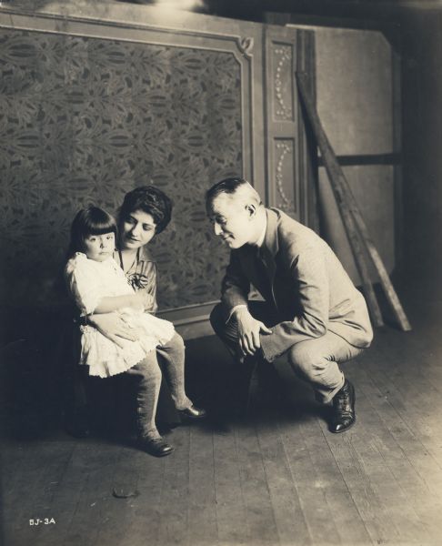 Silent film actress Marguerite Snow with her daughter Julie Jane Cruze on her knee. Behind them are theatrical scenery flats. The older man admiring the child is unidentified.