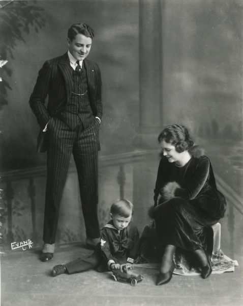 The silent film star Bryant Washburn and his wife the actress Mabel Forrest watch as their little son Bryant Washburn, Jr., plays with a wind-up toy truck on the floor of the Evans photographic studio in Los Angeles. Washburn wears a pin-stripe suit and spats, Forrest wears a black velvet dress with feather cuffs, and the boy wears a sailor suit.