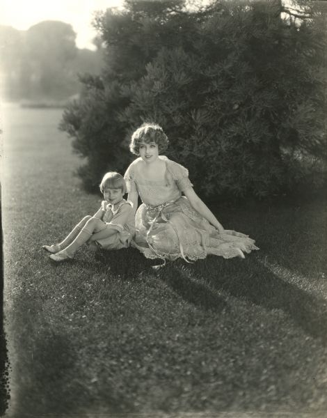 Silent film star Claire Windsor in a lacy summer dress and sun hat sits on the grass with her small son Billy.