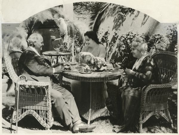 Silent film star Clara Kimball Young, center, with her parents Edward M. Kimball and Pauline Maddern Kimball drinking coffee or tea outdoors. They are seated in wicker chairs around a wicker table, and a cockatoo watches from its perch. Both parents were actors.