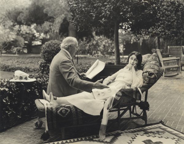 The actor Edward M. Kimball, wearing pince-nez, reads a script to his daughter, the silent film star Clara Kimball Young. She is on a chaise lounge with a magazine in her lap and a far-away expression on her face.