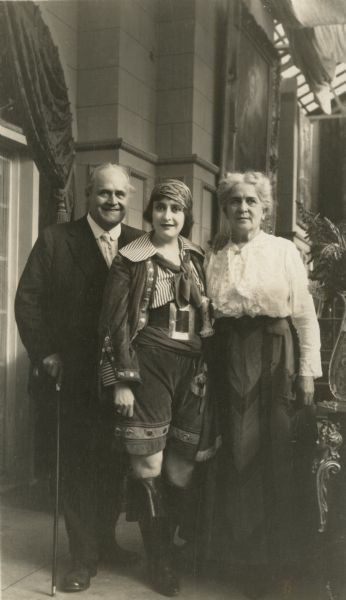 Silent film star Clara Kimball Young, dressed in a pirate costume, with her parents Edward M. Kimball and Pauline Maddern Kimball on either side of her. The skylight in the upper right corner suggests that they are in a motion picture studio.
