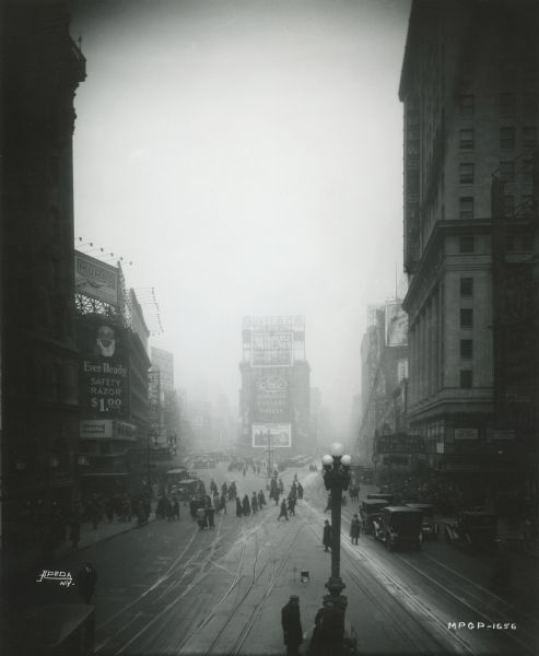Times Square in New York City facing north on a foggy winter day in January or February. On the right is Loew's State Theatre which is playing "Big Brother," a silent film from a story by Rex Beach. Beyond it, another theater is showing "The Hunchback of Notre Dame" with Lon Chaney. On the left at the Astor Theatre, Constance Binney appears in "Sweet Little Devil," a George Gershwin musical.