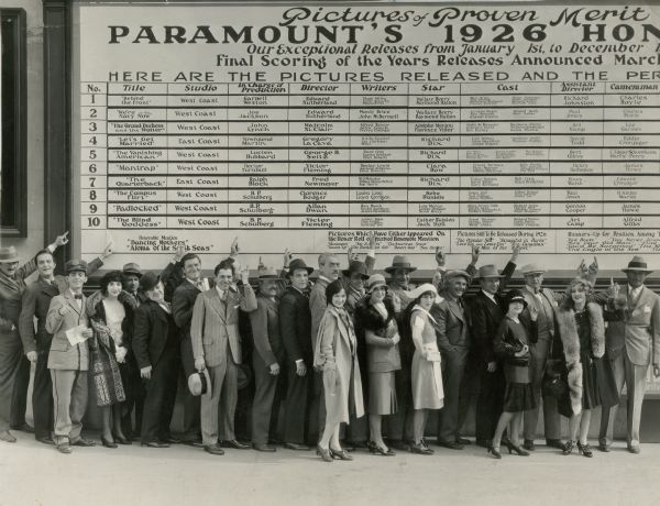 Original caption: "'That's where our names will be next time,'is the solemn vow of this group of Paramount players and directors before the company's 1926 Honor Roll Board. From left to right--John Waters, director, Douglas Gilmore, Eddie Cantor, Bebe Daniels, Jimmy Speak, Assistant Director, Chester Conklin, George Bancroft, Warner Baxter, William Powell, Henri Menjou, Lawrence Gray, Mauritz Stiller, Doris Hill, James Hall, Florence Vidor, Noah Beery, Jobyna Ralston, Clarence Badger, director, El Brendel, Betty Bronson, Clive Brook, Lotus Thompson and Victor Fleming, Director."