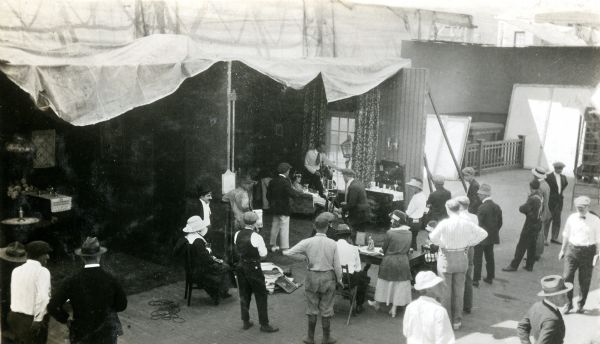 From a high angle, this snapshot shows actors, crew, and bystanders at an open-air studio under canvas diffusers at Universal City. The cameraman stands beside a Bell & Howell motion picture camera. This may be a production of the L-KO Kompany featuring Hank Mann and Katherine Griffith.