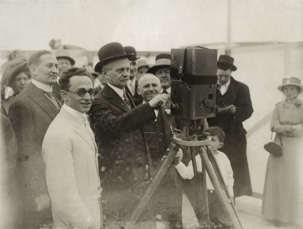 At the grand opening of Universal City are the general manager of the Universal Film Manufacturing Company, Isadore Bernstein (in a white belted Norfolk suit and round sunglasses); the company's president, Carl Laemmle (smiling, between the camera operator and the camera); and, mostly hidden by the tripod, the seven-year-old Carl Laemmle, Jr., his hand held by his father. An unidentified man has his hand on the crank of the Prevost motion picture camera.