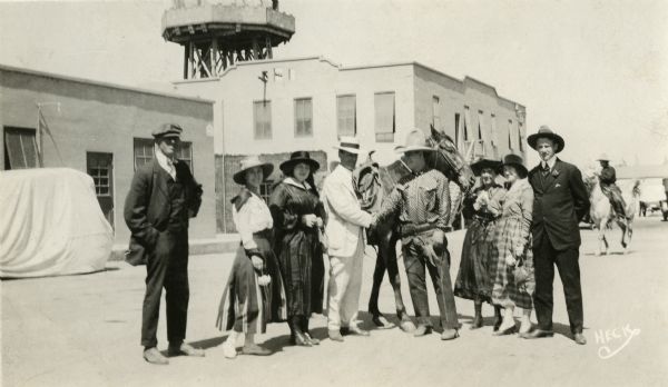 Seven well-dressed tourists visit Universal City and pose with actor Neal Hart who is in cowboy costume and has his horse nearby. A water tower is visible behind them.