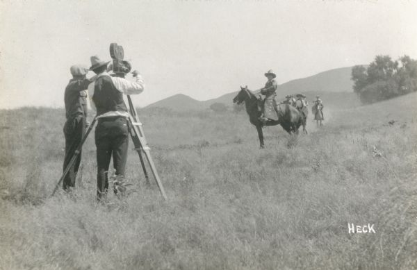 Actor Neal Hart in cowboy costume on a horse being filmed with a Bell & Howell 2709 motion picture camera in the countryside near Universal City. A group of five or six horsemen are galloping behind him.