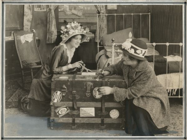 Nellie Nolan (left, played by Edith Taliaferro) and her friend Lou (Florence Dagmar) conspire to make Nellie appear to be the well-traveled heiress Ethel Van Dusen by pasting exotic hotel labels on her steamer trunk. This is a scene still from the silent film "Young Romance" (1915).