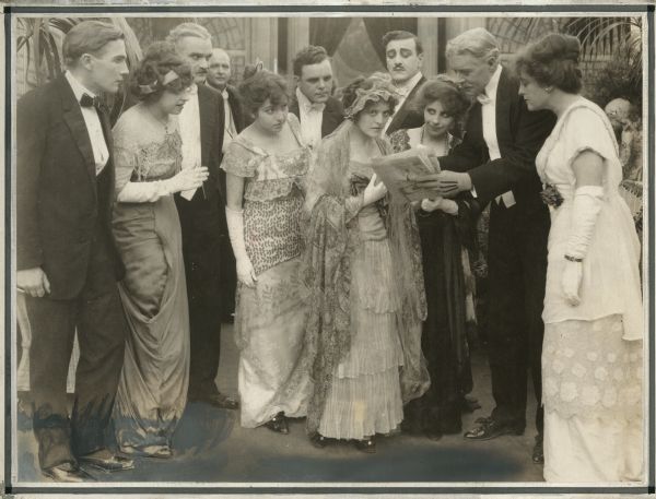 Surrounded by wealthy hotel guests dressed in formal evening wear, Nellie Nolan (played by Edith Taliaferro) learns that the person she is pretending to be has just inherited a fortune. Behind her, the criminal Spagnoli (played by Al Ernest Garcia wearing a small dark moustache) begins to plot.