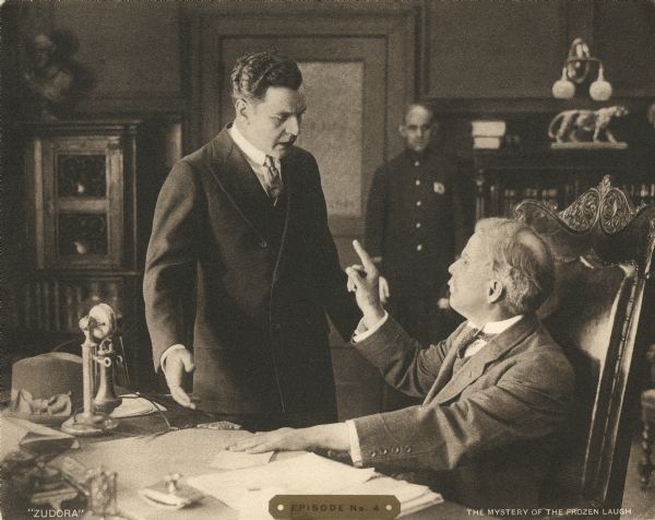 John Storm (played by Harry Benham) is in a heated discussion with a police chief or some other authority (played by Morgan Jones) in a scene still from the Thanhouser serial "Zudora." A police officer stands at the rear of the office.