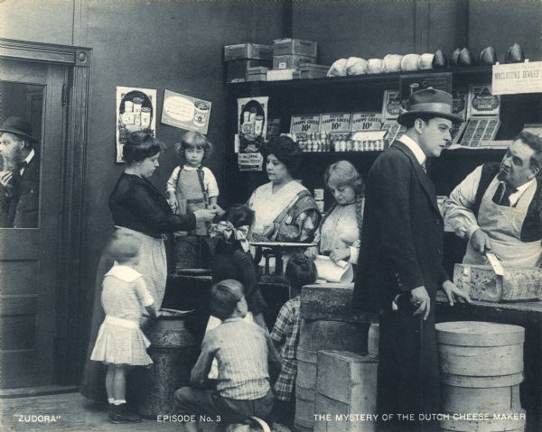 John Storm (played by Harry Benham) talks to a cheese maker (Justus D. Barnes) in his shop in a scene still from the Thanhouser serial "Zudora." The print is marked "Episode No. 3" and "The Mystery of the Dutch Cheese Maker."