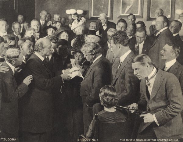 A crowd is gathered in a courtroom in this scene still from the Thanhouser serial "Zudora." The blonde man on the far left is the actor N.Z. Wood. Zudora (played by Marguerite Snow) sits with her back to the camera while Tom Hunt (a detective, played by Sidney Bracey) points a pistol with a silencer at her head. John Storm (played by Harry Benham) is in the striped suit. The print is captioned "Episode No. 1" and "The Mystic Message of the Spotted Collar."