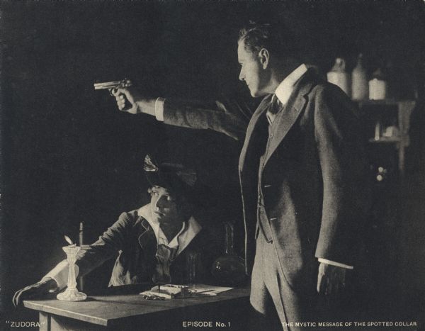 Zudora (played by Marguerite Snow) sits at a table while John Storm (Harry Benham) practices with a semi-automatic pistol in a candlelit basement in a scene still for the Thanhouser serial "Zudora." The print is captioned "Episode No. 1" and "The Mystic Message of the Spotted Collar."