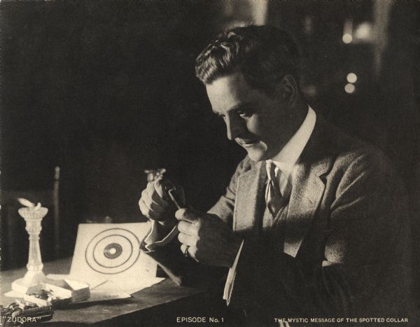 In a candlelit basement, John Storm (played by Harry Benham) loads bullets into a pistol magazine in a scene still for the Thanhouser serial "Zudora." The print is captioned "Episode No. 1" and "The Mystic Message of the Spotted Collar."