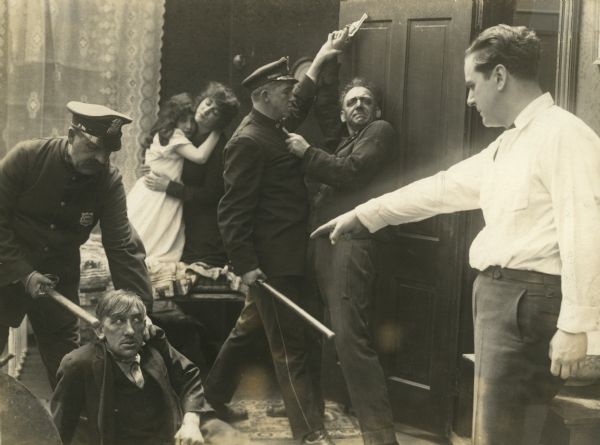 Actor Harry Benham points accusingly at a ruffian on the floor as two police officers subdue two criminals in a scene still from the Thanhouser serial "Zudora." Toward the rear, two women cower in terror.