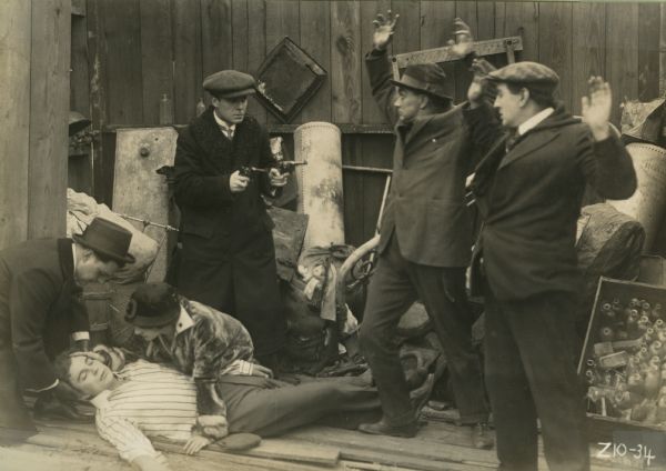 Actor Harry Benham covers two ruffians with two pistols as Marguerite Snow ministers to an insensible young man on the floor in a scene still from the Thanhouser serial "Zudora."