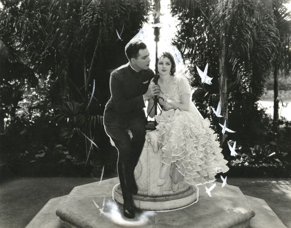Lieutenant Eric Burgess, a U.S. Army aviator played by Harrison Ford, sits on the edge of a well with Marty Mackenzie (Vivian Martin) in a scene still from "You Never Saw Such a Girl" (1919). She wears a white ruffled party dress and he is in military uniform.
