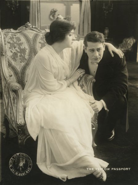 Sonia Sokoloff (played by Clara Kimball Young) in a flowing white gown is seated while Adolph Rosenheimer (Edwin August) is on one knee hanging onto the arm of her chair looking distressed in a scene still from "The Yellow Passport" (1916).