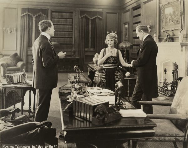 Margaret Vane (played by Norma Talmadge) is the center of attention in a scene still from "Yes or No." On the left, in front of a desk, is Dr. Malloy (played by Lionel Adams), while holding Margaret Vane's hand is her husband, Donald Vane (Frederick Burton). They are in a richly decorated private office with bookcases and a fireplace.