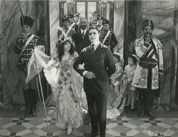 Princess Alexia (played by Enid Markey in an elaborately decorated dress and wearing a crown) marches arm in arm with Dick Mason (George Walsh) in a scene still from the 1917 Fox production "The Yankee Way." Directly behind them is the bearded Baron Maravitch (Count Fritz von Hardenberg).