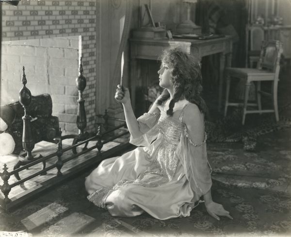 Princess Pat (played by Gladys Leslie) sits by the fireplace and examines her face in a hand mirror in a scene still for "The Wooing of Princess Pat."