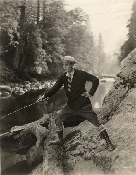 In a publicity still used to promote "The World's Champion," the silent film star Wallace Reid is shown fishing in a river, probably in Yosemite National Park. Reid wears a cloth cap, cardigan sweater, striped tie, jodhpurs, high boots, and white pancake makeup.