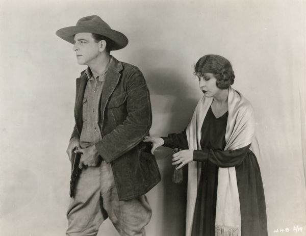 Iris Champneys (played by Dorothy Dalton) comes up behind Clement Gaunt (Milton Sills) apparently to sneak a small money bag into the pocket of his corduroy jacket in a publicity still for "The Woman Who Walked Alone." Sills is costumed in western wear including jodhpurs and a campaign hat.