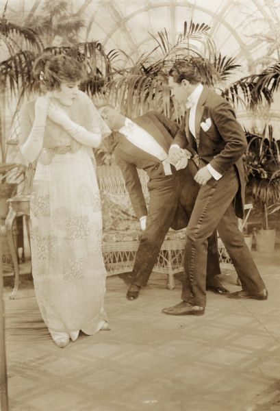 Ethel Clayton in a floor-length Japanese print dress and long white gloves flinches as Joseph Kaufman punches an unidentified actor in a scene still from "A Woman Went Forth" (Lublin 1915). Both men are wearing white ties and tailcoats. They are in a conservatory with wicker furniture and potted palms.