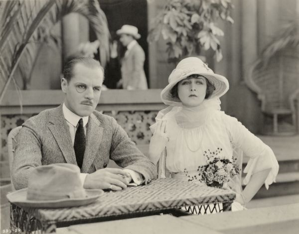 Lord Raa (played by Jack Holt wearing a monocle and a mean look) sits beside Mary MacNeill (Katherine MacDonald in a white dress and hat) in a scene still for "The Woman Thou Gavest Me."