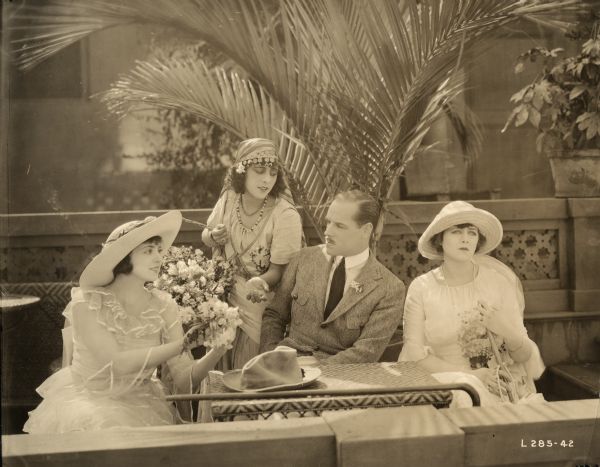 Lord Raa (played by Jack Holt wearing a monocle) sits beside Mary MacNeill (Katherine MacDonald in a white dress and hat) who is looking miserable in a scene still for "The Woman Thou Gavest Me." To the left are a bridesmaid and a Gypsy girl selling flowers played by unidentifed actresses.	