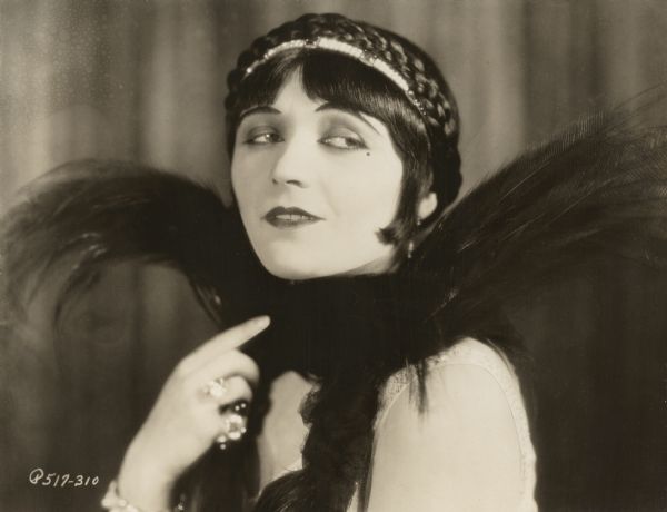 A glamorous head and shoulders publicity still of Pola Negri dressed for the role of the Countess Elnora Natatorini in the 1925 Paramount production "A Woman of the World." She wears a jeweled headband, large rings, and black feathers.