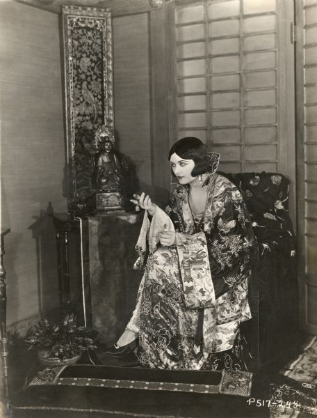 This glamorous scene still shows Pola Negri dressed for the role of the Countess Elnora Natatorini in the 1925 Paramount production "A Woman of the World." She sits in front of a Buddha and wears an elaborate Chinese silk robe with a high collar of black lace outlined in pearls.