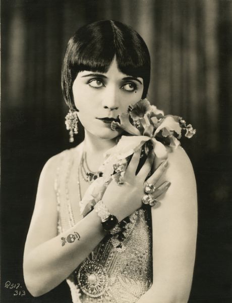 This glamorous half-length publicity still shows Pola Negri dressed for the role of the Countess Elnora Natatorini in the 1925 Paramount production "A Woman of the World." She is wearing a dress encrusted with pearls and jewels, and has her cheek against an orchid she is holding up on her shoulder. On her right forearm is a tattoo of a butterfly and a skull.