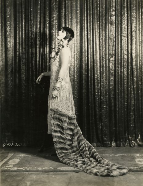 Pola Negri stands in front of a shiny satin theater curtain and is dressed for the role of the Countess Elnora Natatorini in the 1925 Paramount production "A Woman of the World." Her dress is encrusted with pearls and jewels ends in a train made of fur. On her shoulder is an orchid.