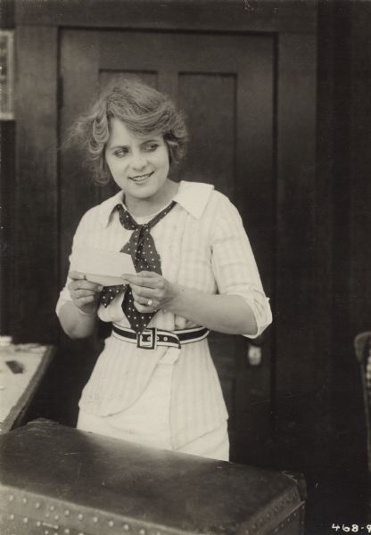 Kathlyn Williams grins and holds a letter in this three-quarter length publicity still for the 1914 Selig production "A Woman Laughs." She stands behind a suitcase.
