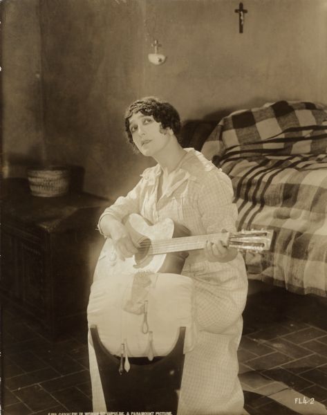 Leonora (played by Lina Cavalieri) is the daughter of a poor Italian lacemaker in this scene still from "A Woman of Impulse." She strums a guitar in an impoverished bedroom. In the foreground is a lacemaker's pillow on a stand with bobbins hanging from a piece of lace. On the wall behind her are a small crucifix and a holy water font.