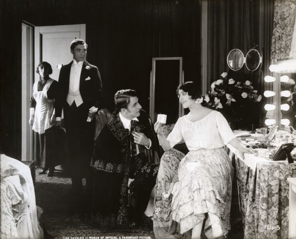 In a scene still for the silent drama "A Woman of Impulse," Leonora, the opera singer known as "La Vecci" (played by Lina Cavalieri) sits in her backstage dressing room receiving visitors. She holds a small gift box that Count Nerval (Raymond Bloomer, on one knee wearing a richly decorated Spanish cape) has just given her. Behind them, looking incensed is Nerval's cousin Phillip Gardiner (Robert Cain, in evening dress). At the door behind him is Leonora's maid.