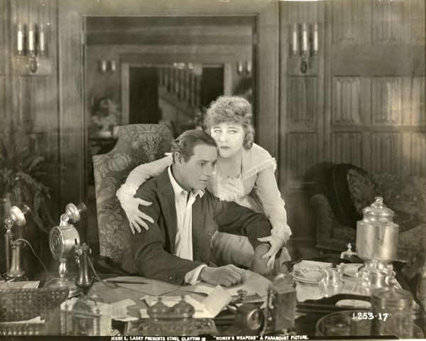 In a scene still for "Women's Weapons," Nicolas Elliot (played by Elliot Dexter) sits at his desk looking stunned. His wife Anne Elliot (Ethel Clayton) sits on the arm of his chair with her arm wrapped around his shoulder. On the desk are a coffee urn and two candlestick telephones. The one on the right appears to be a Strowger Automatic.
