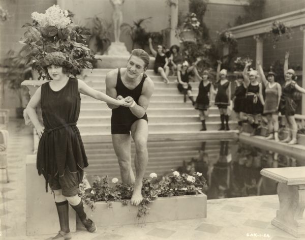 In this flirtatious scene still from "The Wonderman," Henri D'Alour (played by Georges Carpentier, the French boxer, aviator, and actor) holds the hand of Dorothy Stoner (Faire Binney). They wear bathing suits and are standing at the edge of a swimming pool. Behind them a crowd of nine young women also in bathing suits are waving.