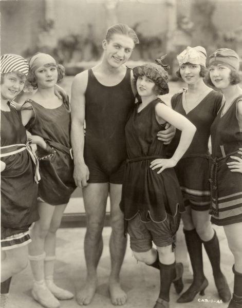 In a publicity still from "The Wonderman," Henri D'Alour (played by Georges Carpentier, the French boxer, aviator, and actor) stands with his arm around Dorothy Stoner (Faire Binney). They wear bathing suits and beside them are four young women also in bathing suits.

