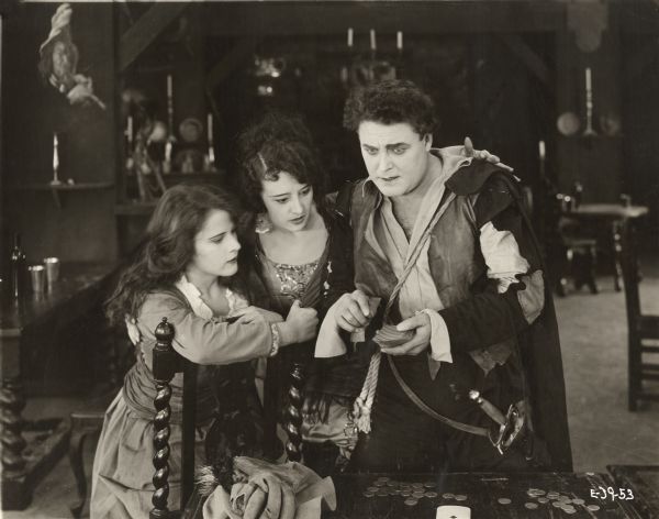 Maritana, a gypsy dancing girl played by Estelle Taylor, has her arm around Don Caesar de Bazan (William Farnum) who holds a deck of cards in a scene still from the 1920 Fox production "The Adventurer." The young actress at left is unidentified.