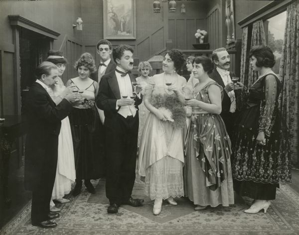 Charlie Chaplin, in tuxedo, surrounded by guests in the party scene from "The Adventurer." He is speaking to Marta Golden who holds a feather fan. In the back, wearing a striped vest is Albert Austin, playing the butler. The small man at the far left is Loyal Underwood.