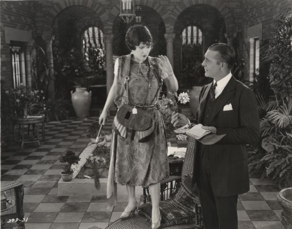 Gloria Swanson stands on a chair with a fishing net in her hand and a fishing basket around her neck as Wallace Reid holds out a flower in a scene still from the Cecil B. DeMille production "The Affairs of Anatol."
