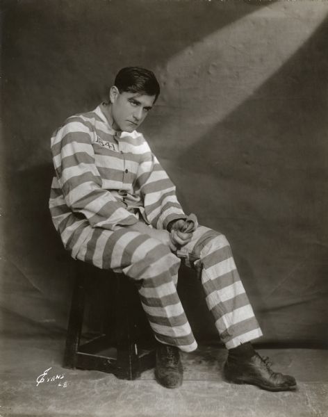 Bert Lytell poses in a prisoner's striped uniform in a publicity still for the silent film "Alias Jimmy Valentine" (Metro 1920). A dramatic shaft of light is on the wall behind him.