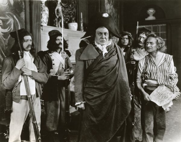 Emil Jannings is costumed as the French Revolutionary leader Georges Danton in a scene still from the silent 1921 production "All for a Woman" (known in Europe as "Danton"). He wears a jacket with very large lapels and on his head is a bicorn. The two soldiers on the left wear Phrygian caps.