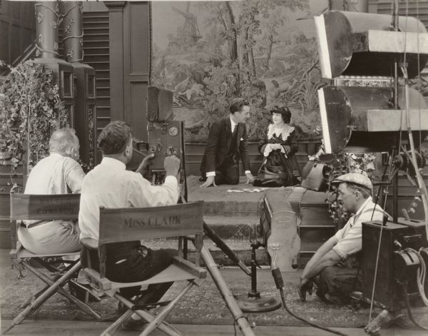 In this production still for "All of a Sudden Peggy," Jack Mulhall and Marguerite Clark kneel and act a scene in front of a tapestry. In the foreground, Walter Edwards sits in his director's chair at left (with a pince-nez stored on his right ear), and next to him cameraman William Marshall cranks a Pathé studio camera. The gaffer sits on the floor at left behind a small Kliegl spotlight and under several Cooper-Hewitt lights.