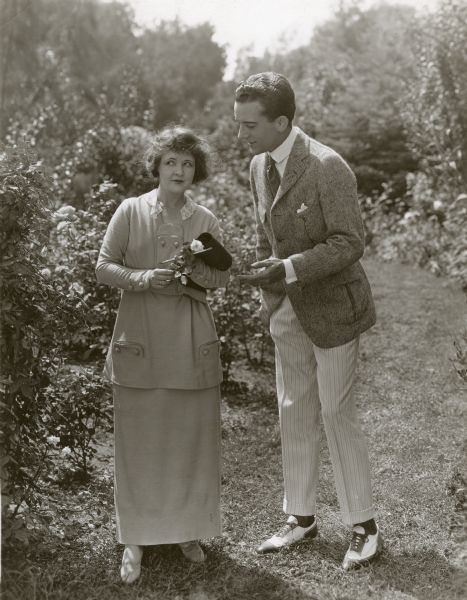 In this publicity still for "All of a Sudden Peggy," Marguerite Clark and Jack Mulhall are talking outdoors in a rose garden. Both are dressed very fashionably. Clark wears a light-colored suit consisting of a tunic over a long, thin skirt. Mulhall wears a jacket of salt and pepper wool with inverted pleats on the pockets, white trousers with pin stripes, and two-toned wingtip shoes.