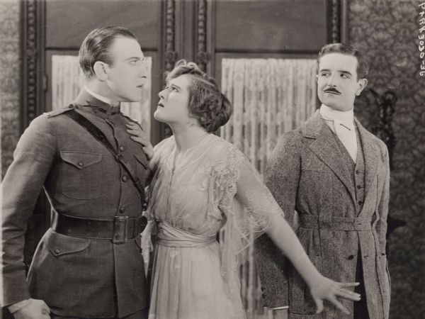 In a scene still from "The Amazing Wife," U.S. Army Lieutenant John Ashton (played by Frank Mayo) clinches his fist and glares at his cousin Philip Ashton (Stanhope Wheaton). Separating them is Cicely Osborne (Mary MacLaren). Mayo wears an army uniform with a Sam Browne belt, and Wheaton is costumed in a belted Norfolk jacket and ascot.